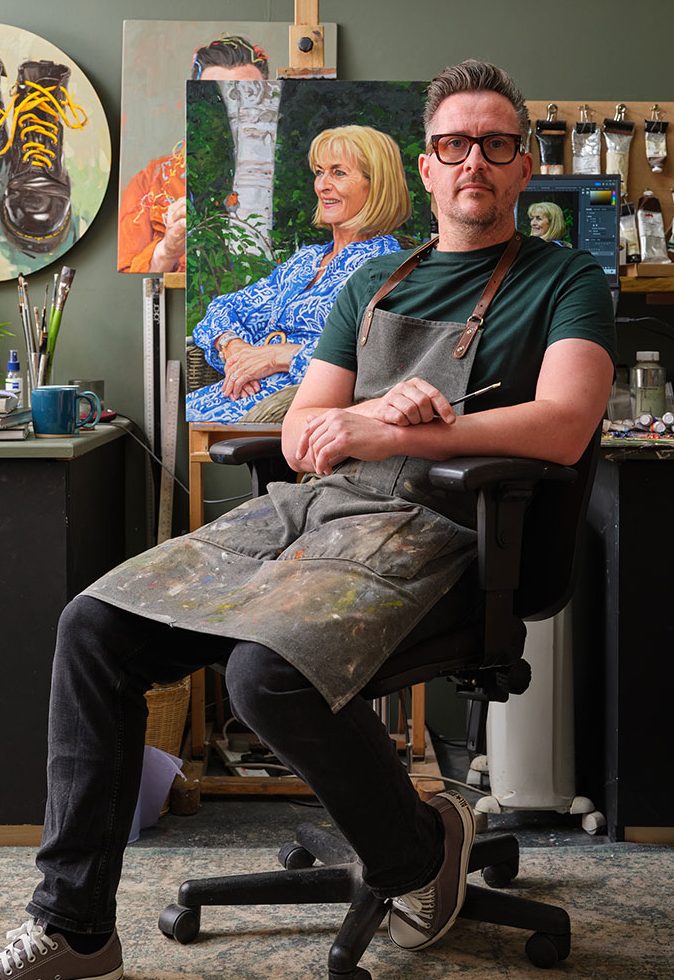 Portrait artist Oliver Winconek sat in his art studio in the UK holding a brush, paintings and art materials are in the background