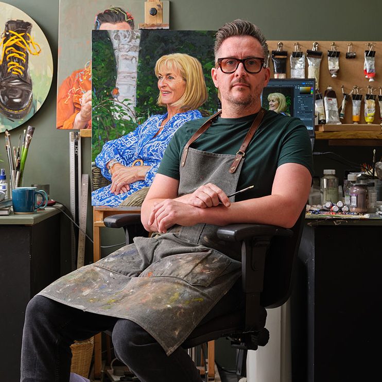Portrait artist Oliver Winconek sat in his art studio in the UK holding a brush, paintings and art materials are in the background
